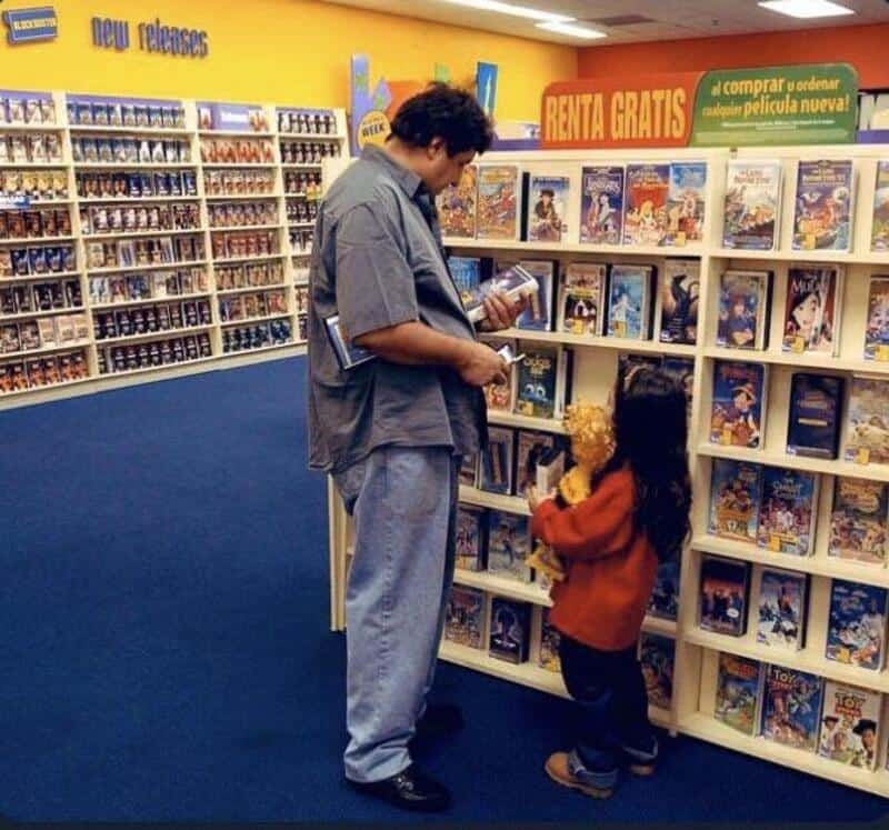 Man and girl browsing comic books in a store, shelves filled with comics and DVDs.