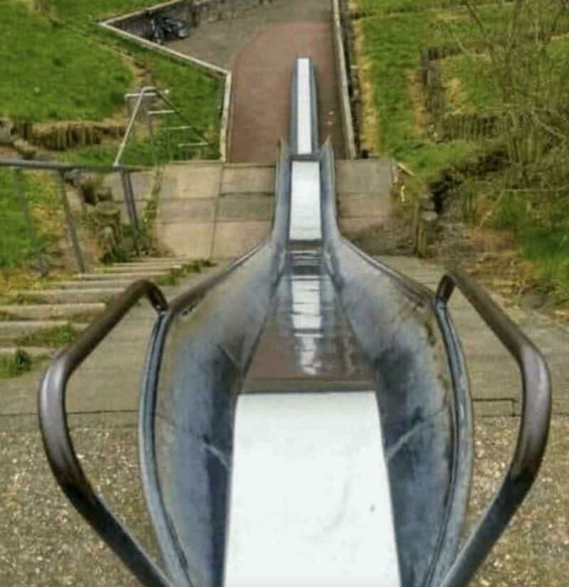 Empty metal slide with stairs and green grass on sides, viewed from top.