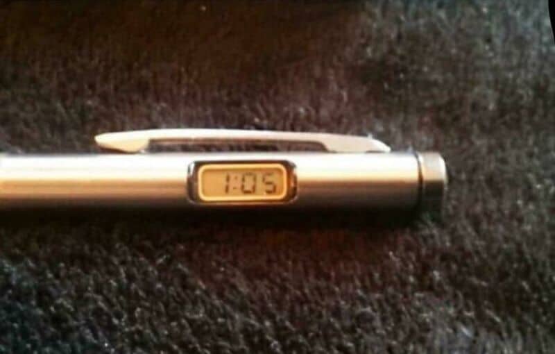 Digital clock on a pen showing 1:05, on a dark textured surface.