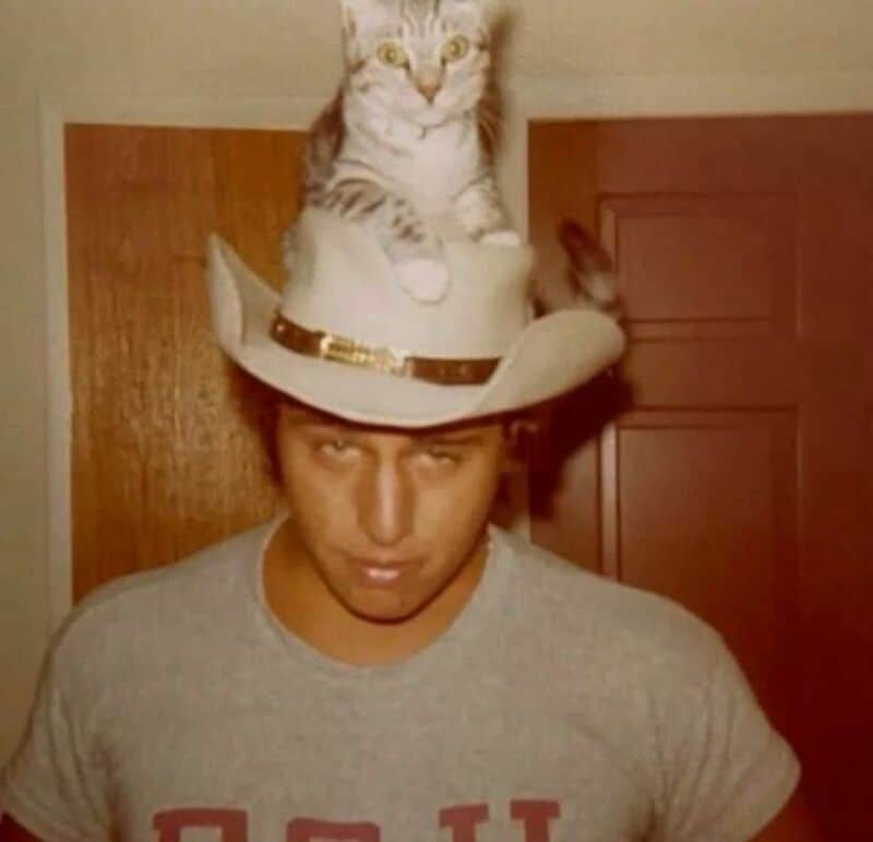 Young man with a cat on his cowboy hat in front of a wooden door.