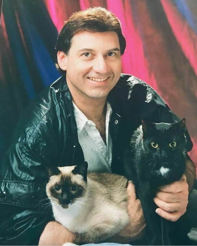 Man with two cats, one Siamese and one black Persian, in front of colorful backdrop.