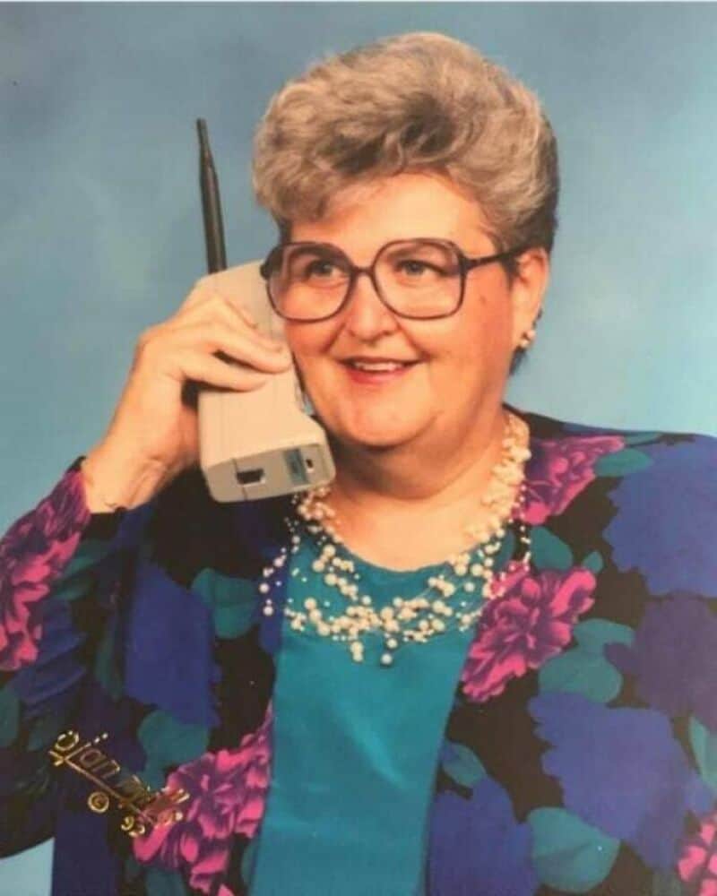 Older woman with glasses talking on a cordless phone, wearing a floral blazer and pearl necklace.