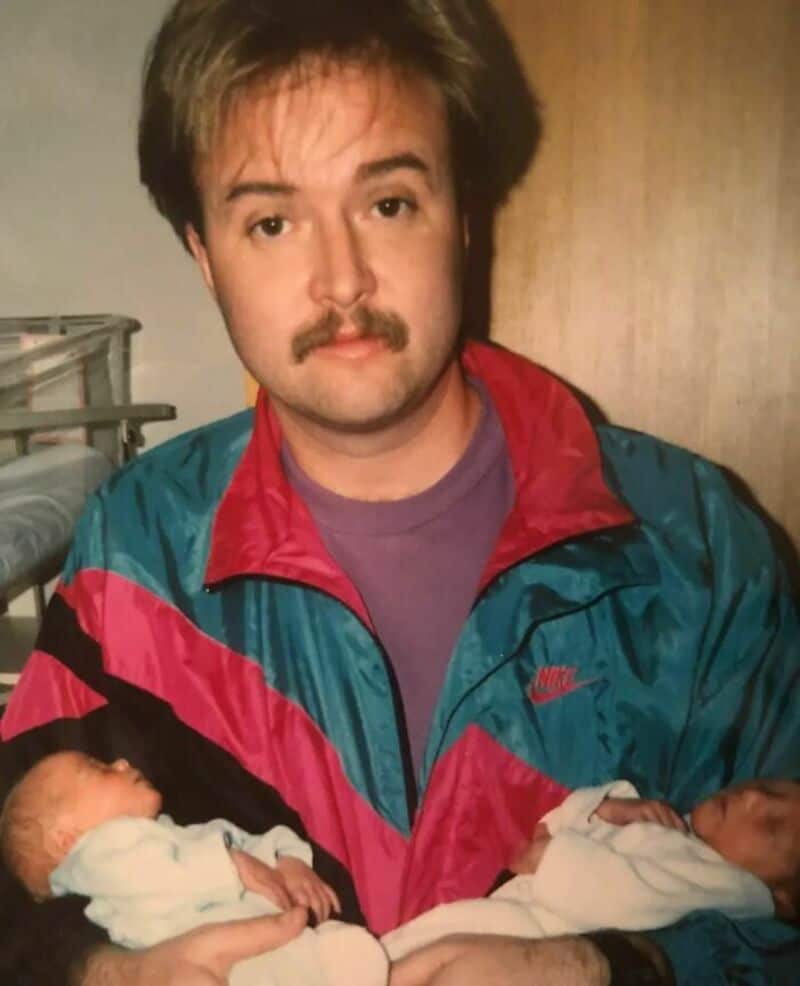 Man with mustache holding two newborn babies, wearing colorful jacket.