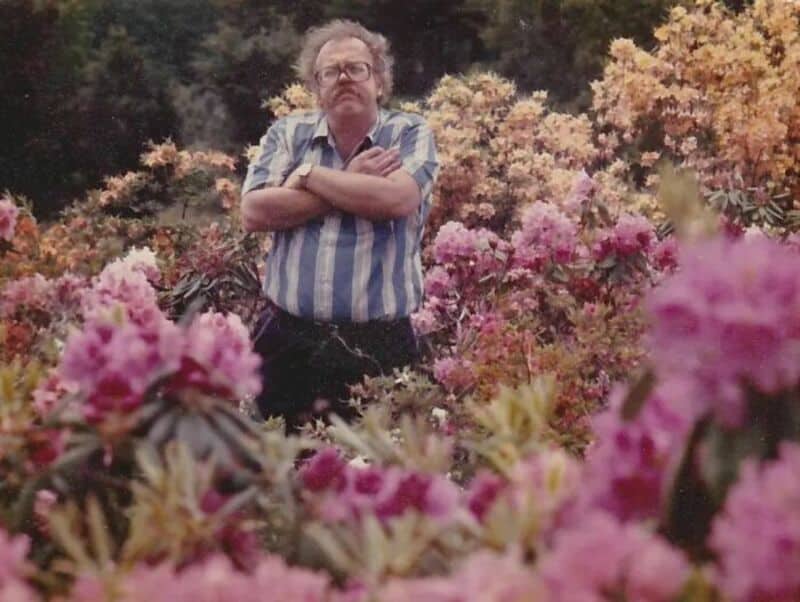 Man with glasses and striped shirt standing among pink and purple flowers in a garden.