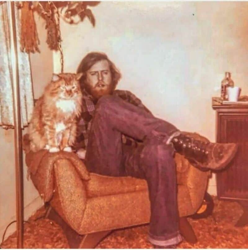 Man with long hair and beard sitting on a couch with a large orange cat.