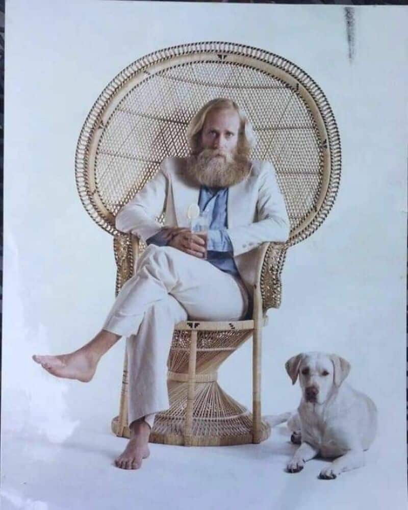 Man with long beard sitting in a wicker chair with a dog beside him.