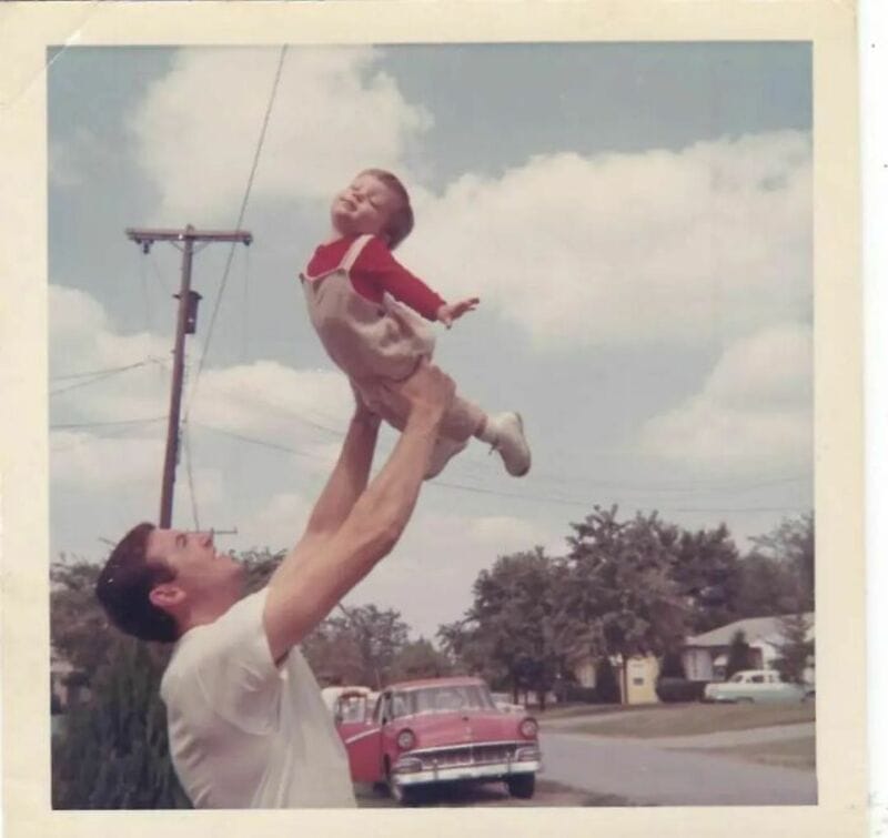 Man lifting child in the air outdoors with a car and houses in background.