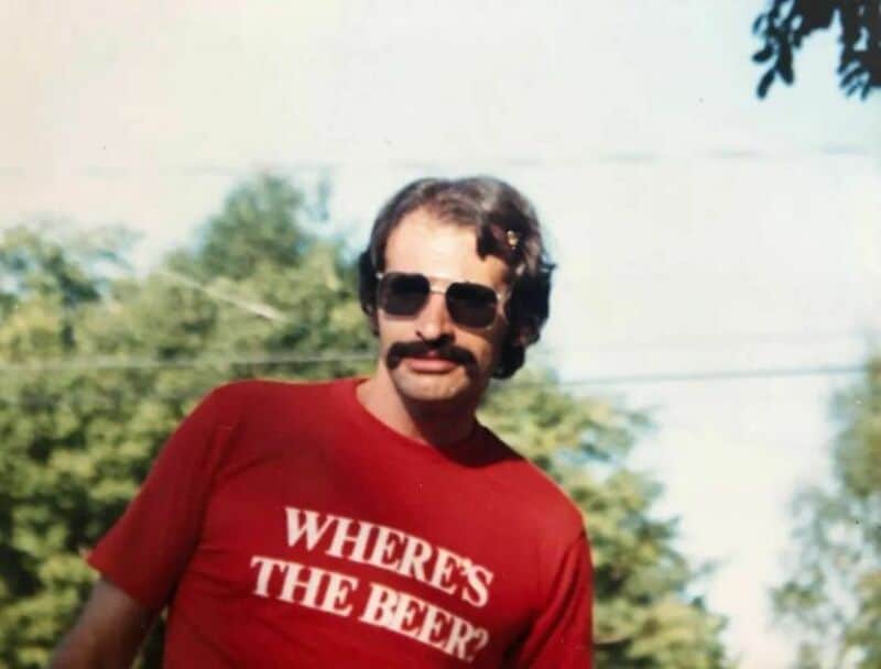 Man with mustache and sunglasses wearing a red T-shirt that says 'WHERE'S THE BEER?'.