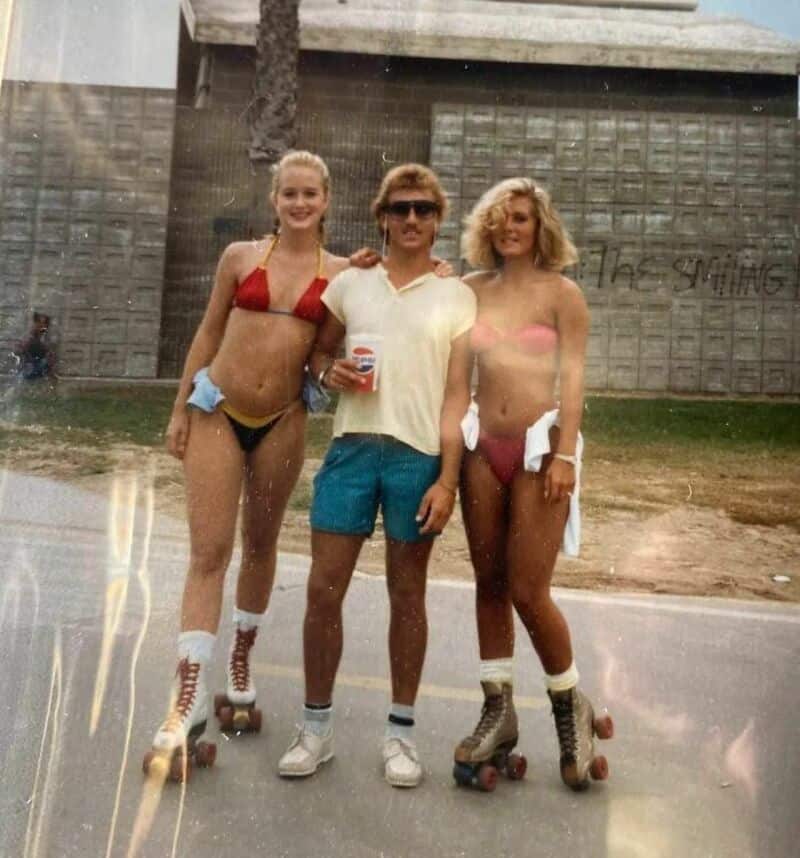 Two women in bikinis and a man in shorts and sunglasses on roller skates outside.