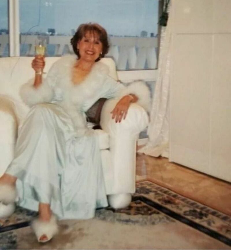 Woman in white dress with fur trim sitting on a white sofa, holding a glass of champagne.