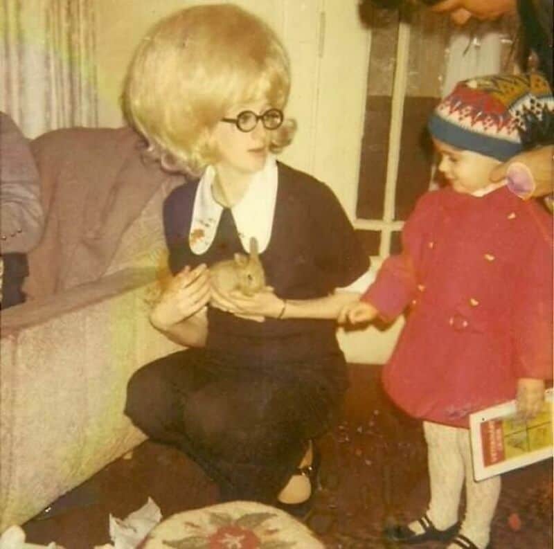 A woman with blonde hair and glasses holding a small animal, with a child in a red coat and colorful.