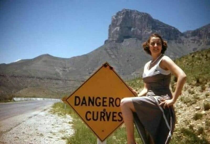 Woman sitting on a rock next to a 'Dangerous Curves' road sign in a mountainous area.