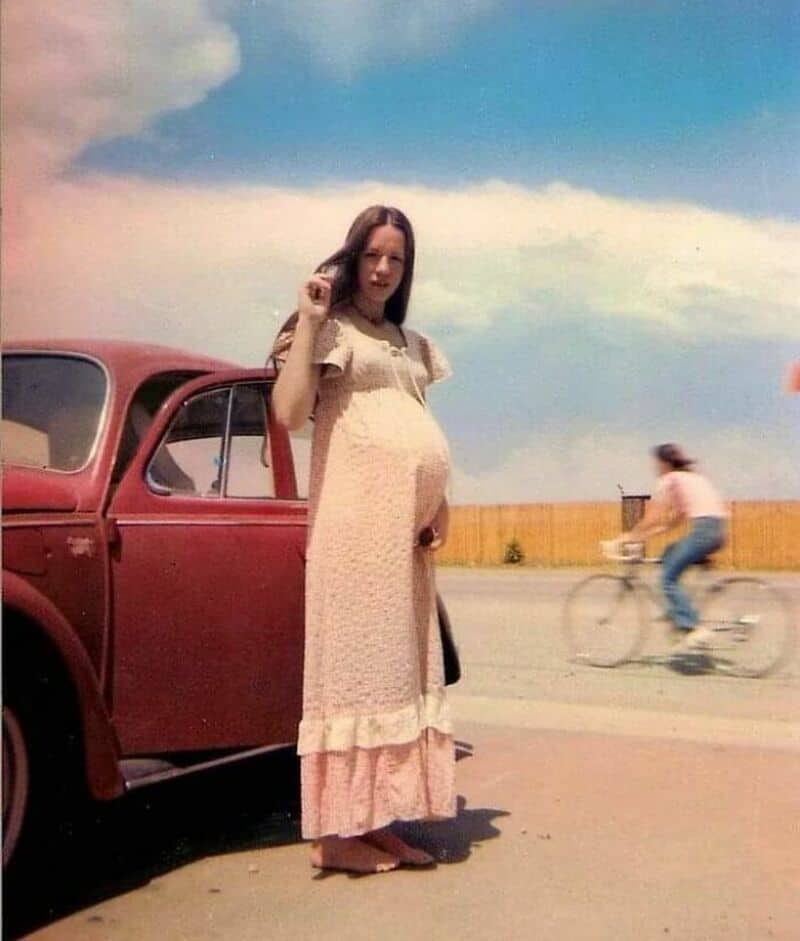 Woman in vintage dress standing next to a red car with a cyclist in the background.