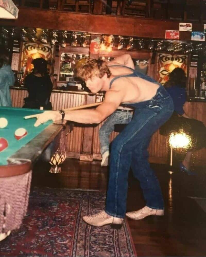 Young man with curly hair playing pool in a bar, wearing a sleeveless top and jeans.