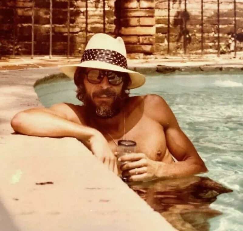 Man with beard, sunglasses, and hat relaxing in pool with drink.