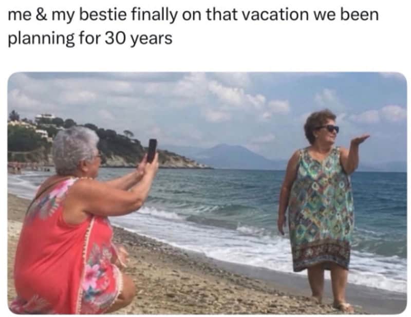 Two women on a beach, one taking a photo of the other, with a scenic ocean view.