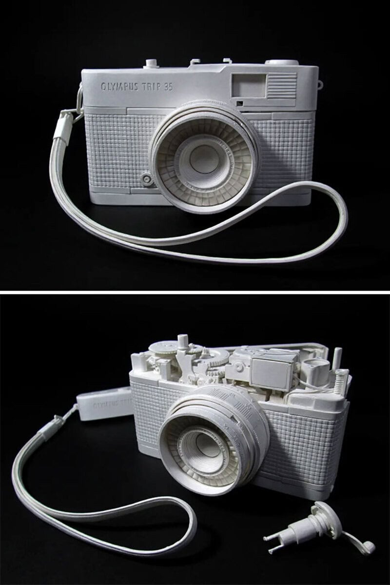 White paper sculpture of a vintage camera with a detachable lens and strap.