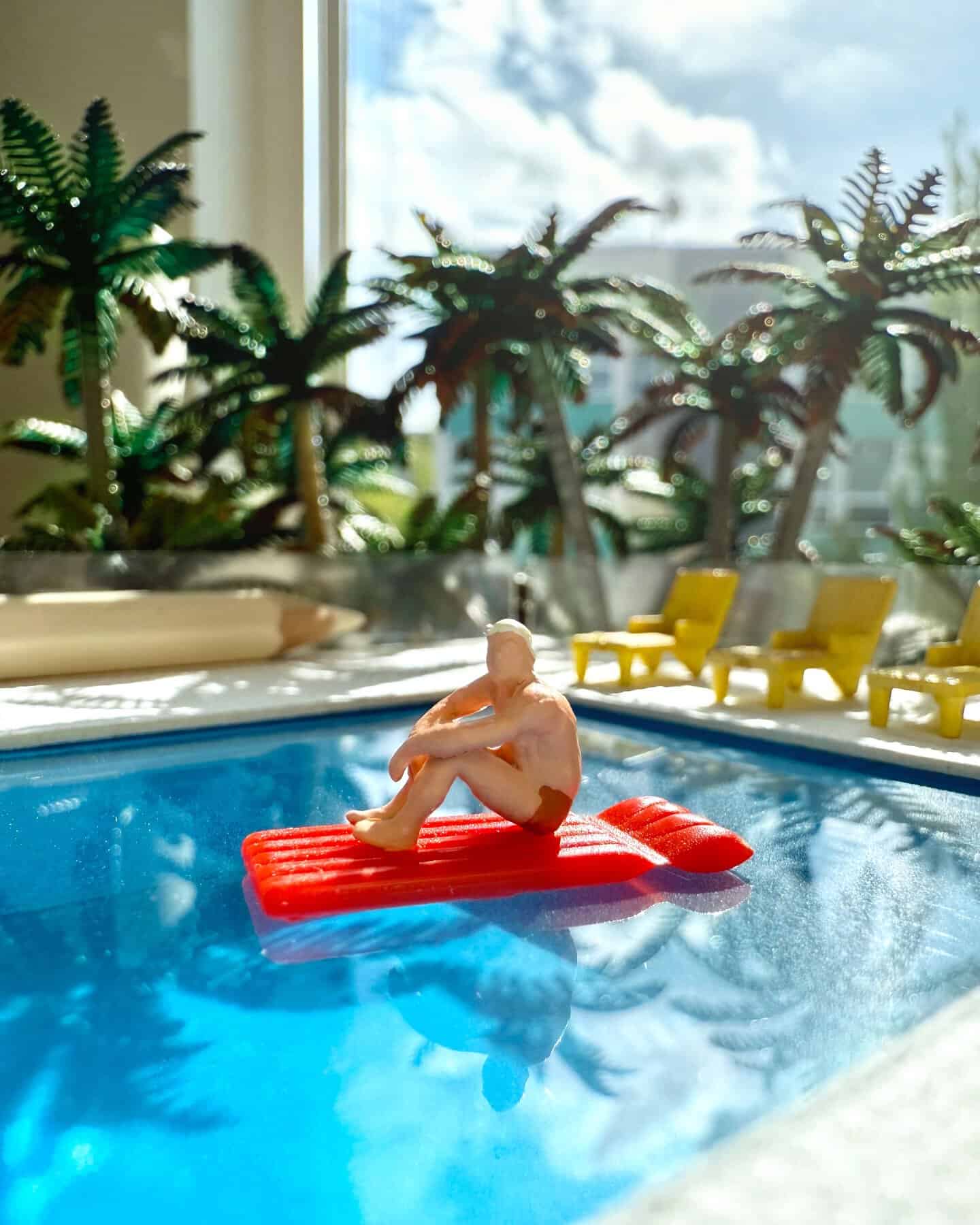 Miniature figurine on a red float in a swimming pool with palm trees outside.