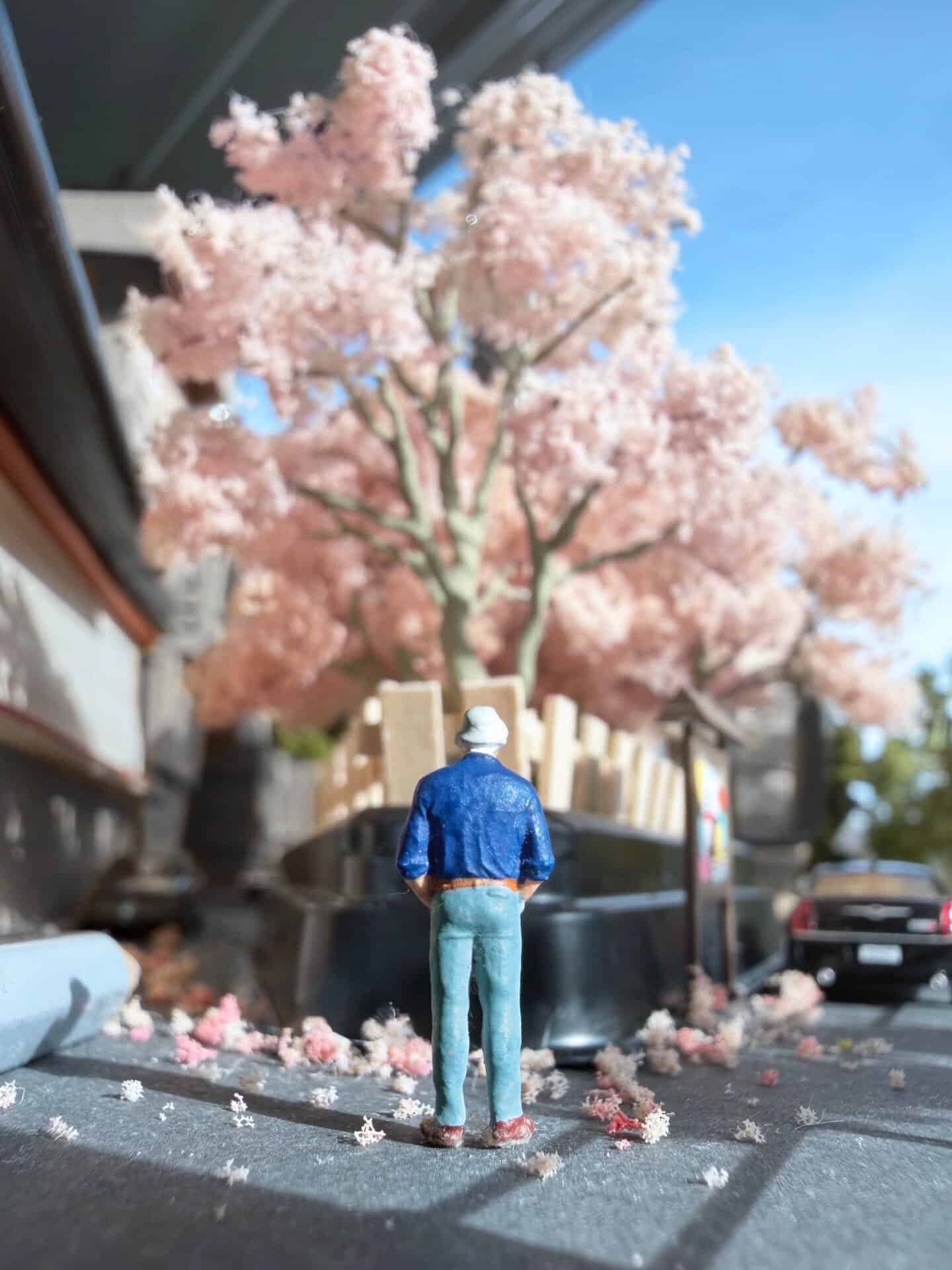 Miniature figure stands in front of a pink cherry blossom tree on a street.