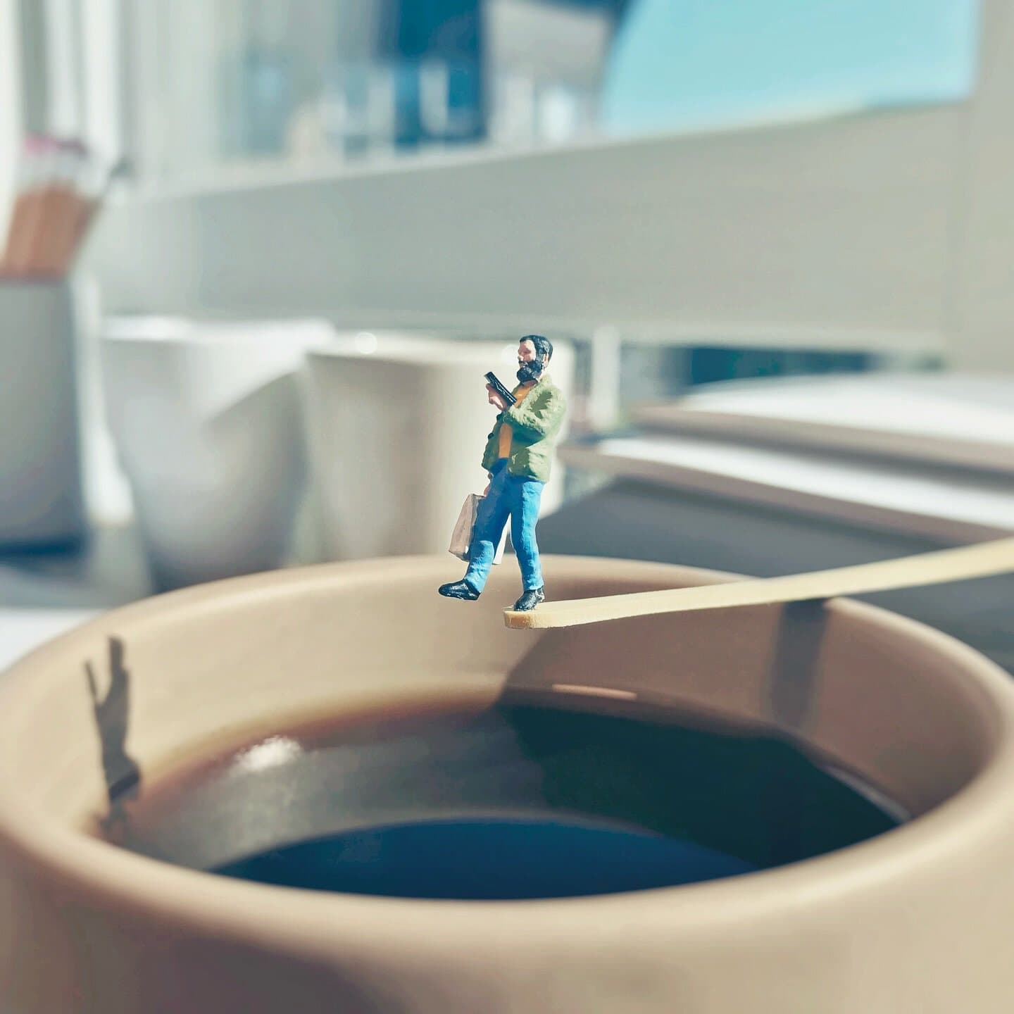 Miniature man figure sitting on the edge of a coffee cup, reading a book.