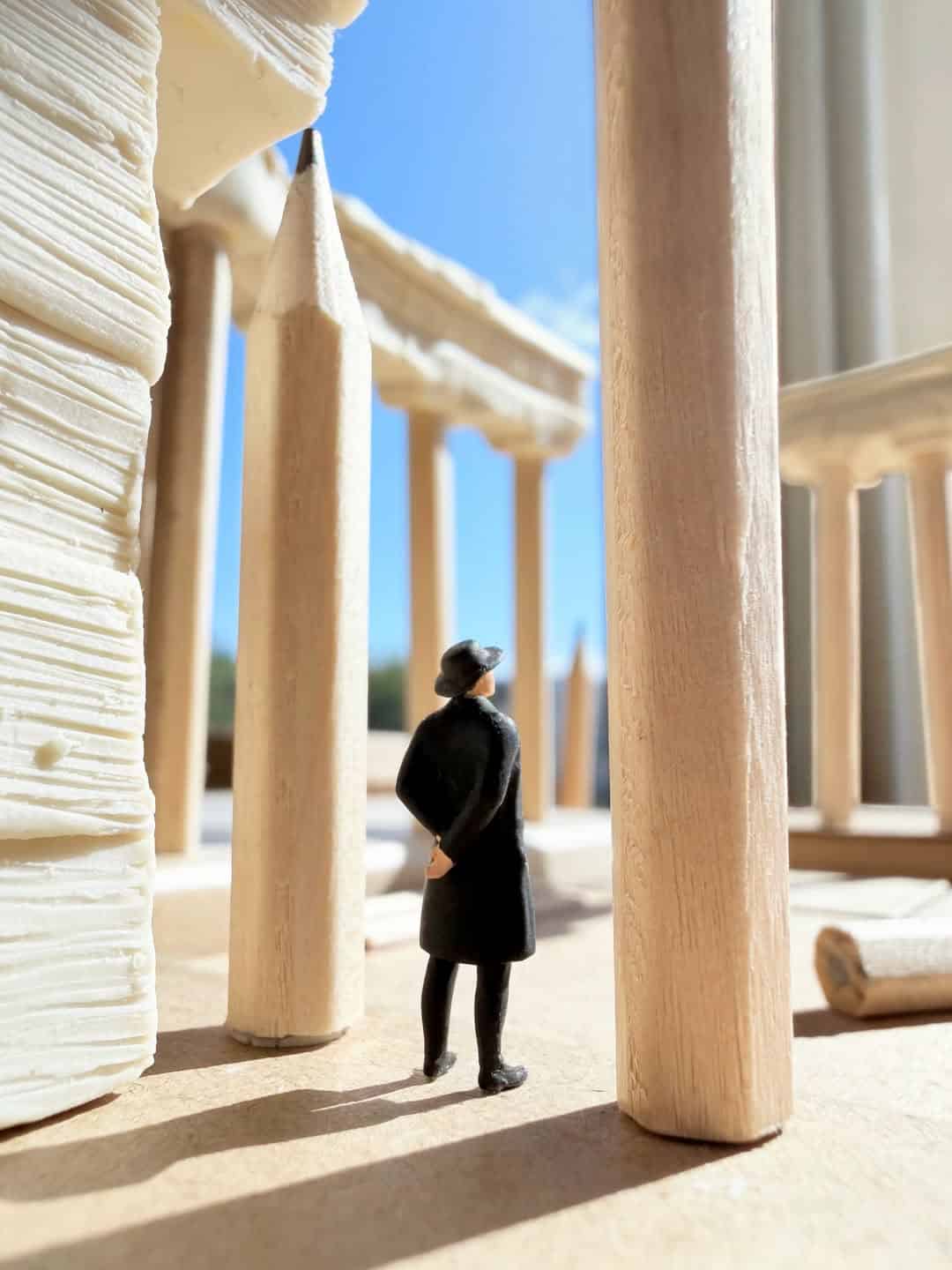 Miniature person in black coat and hat among large wooden structures and paper-like walls.