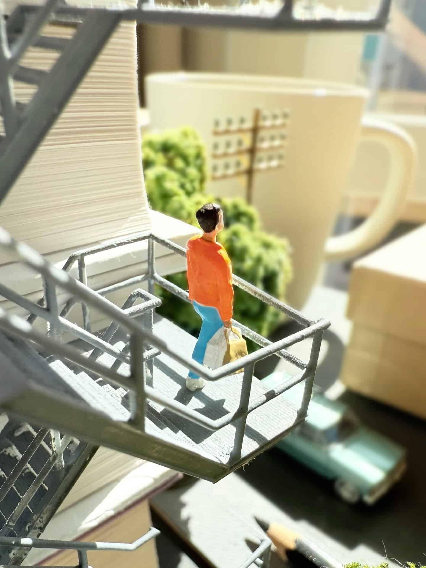 Miniature figure of a person on a balcony with a mug and greenery in the background.