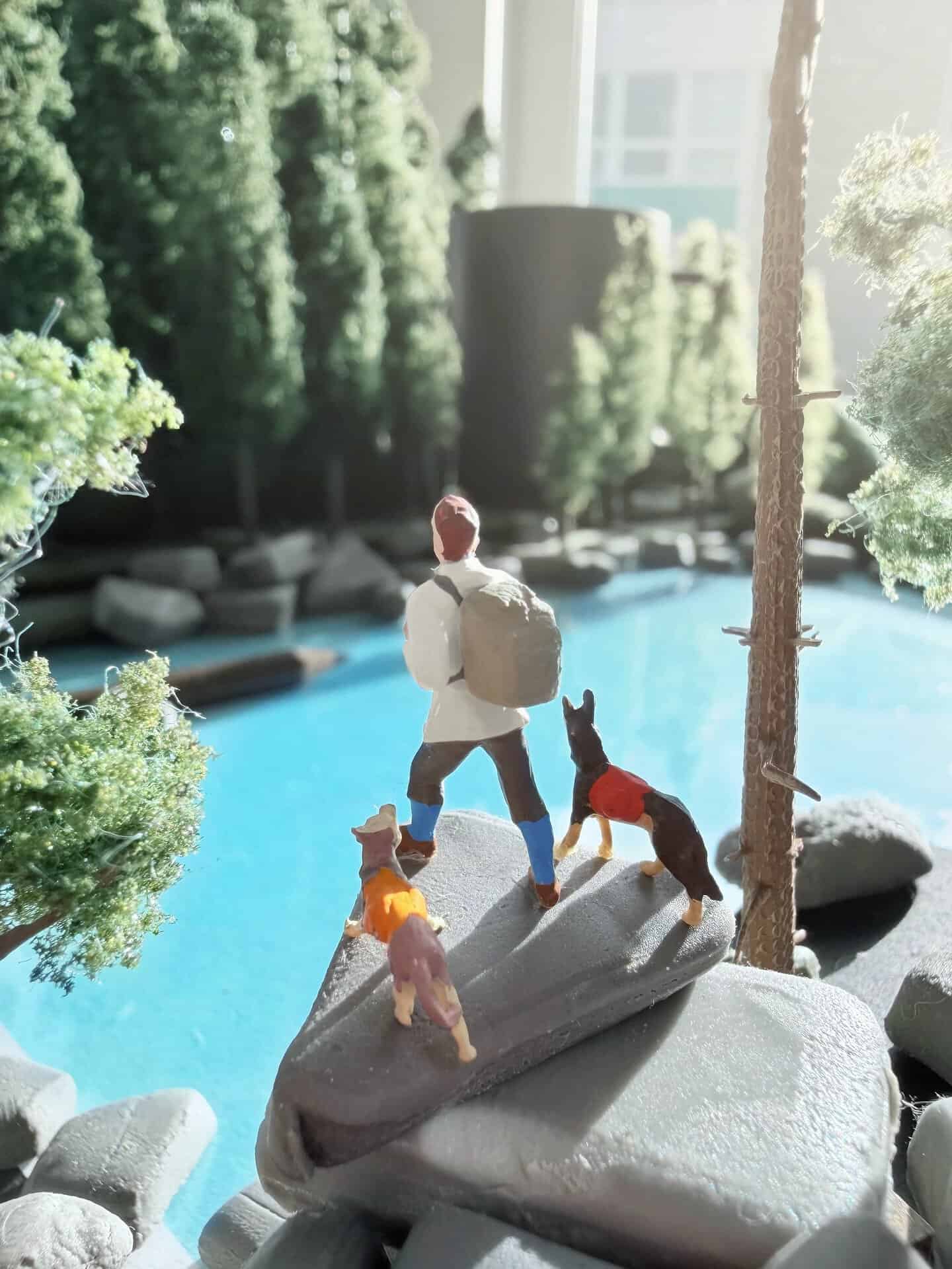 Miniature figure with dog on a rock by a pool, surrounded by trees.