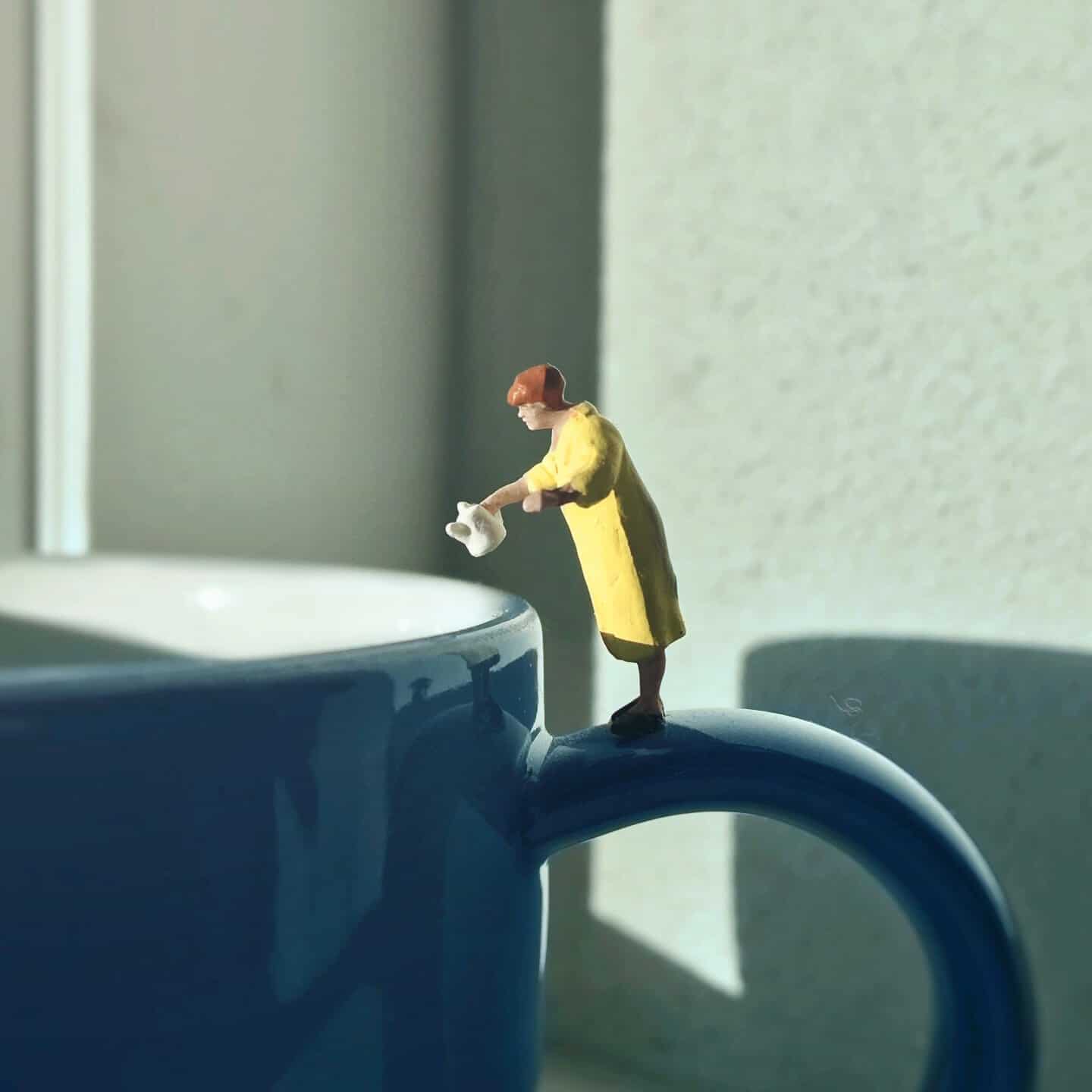 Miniature woman watering a large blue mug with sunlight and shadow effects.