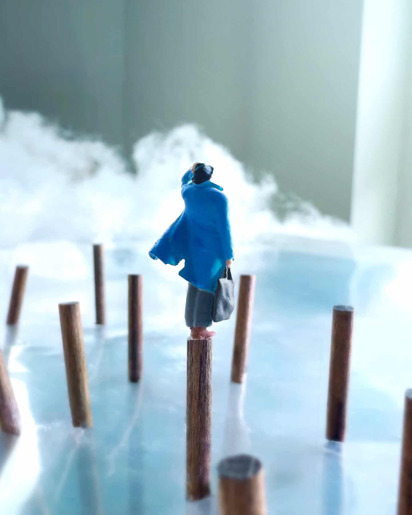 Miniature figure in blue coat stands on a wooden stick among other sticks, with fog and clouds in a.