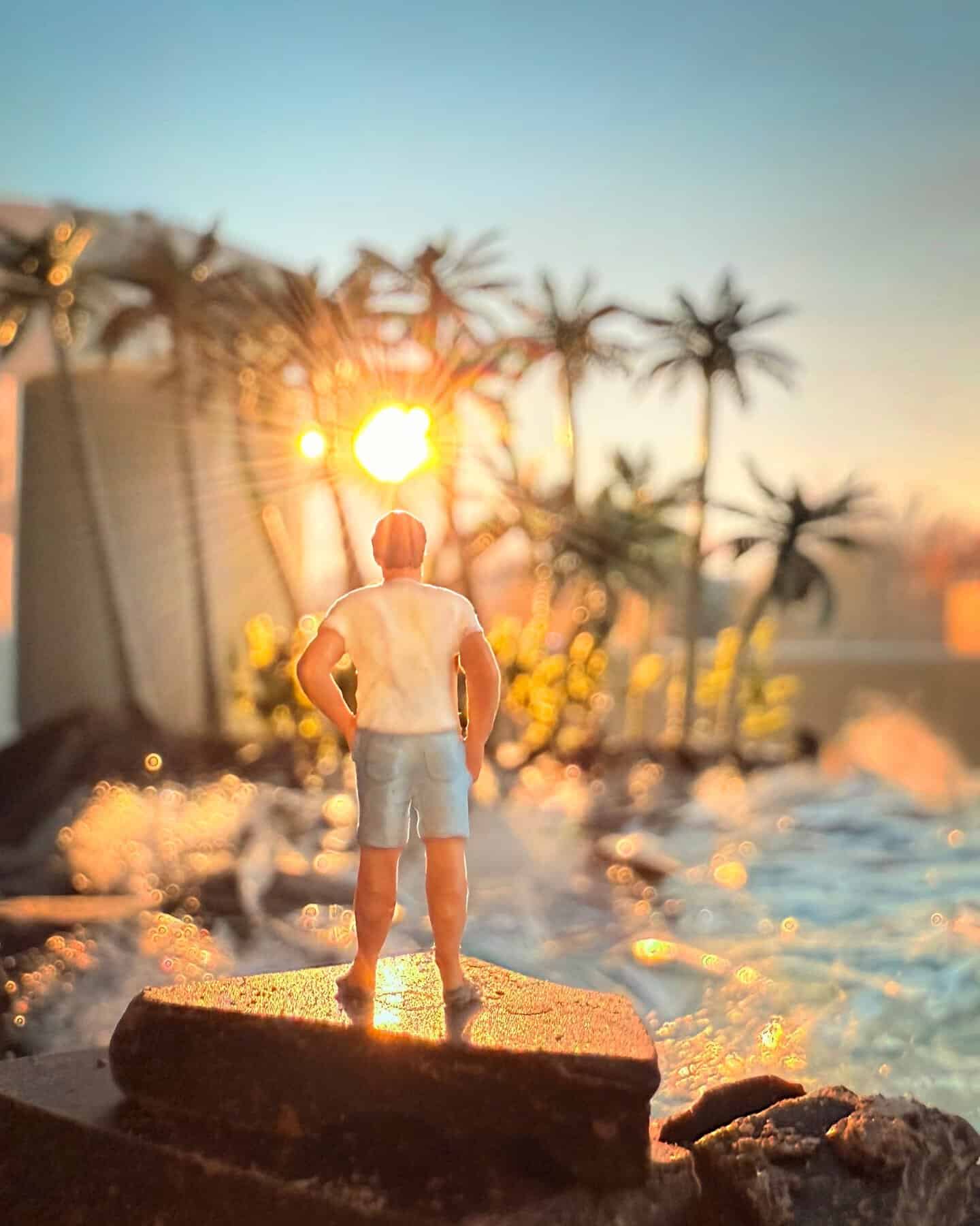 Miniature man on a rock by a pool with palm trees and sunset in background.