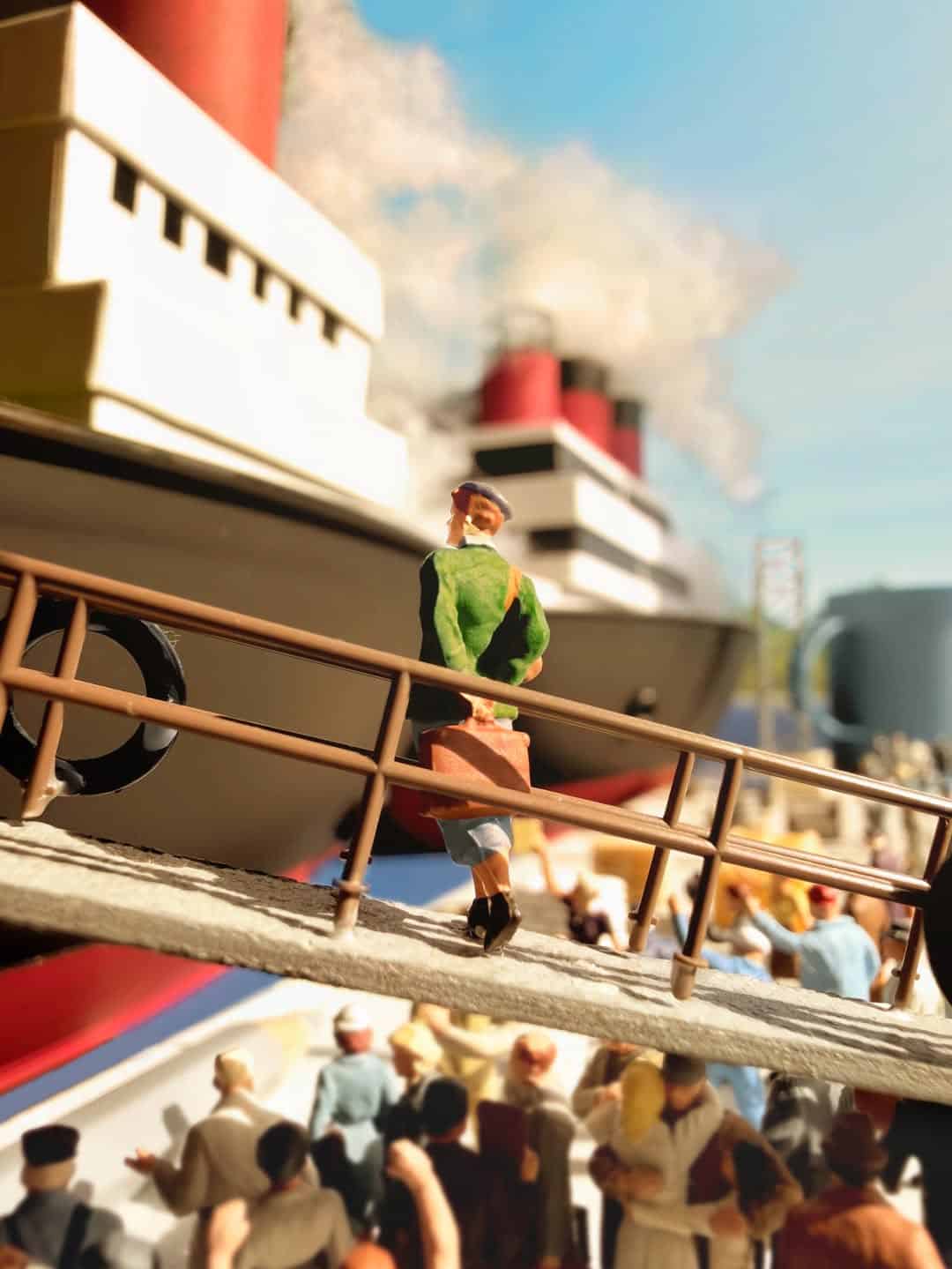 Miniature figure of a man walking on a bridge with a large ship in the background.