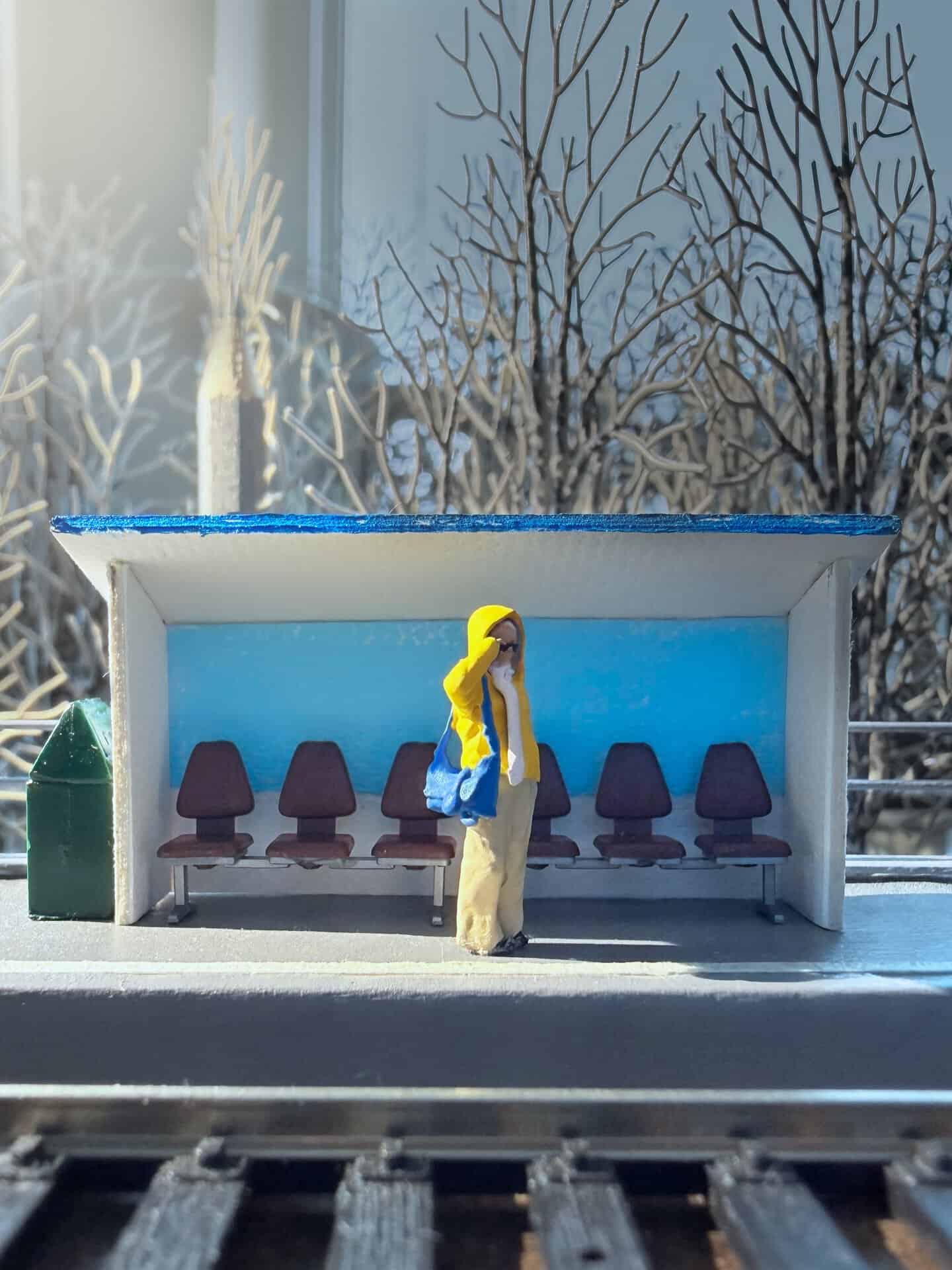 Miniature bus stop with a figure of a woman and snow-covered trees outside.