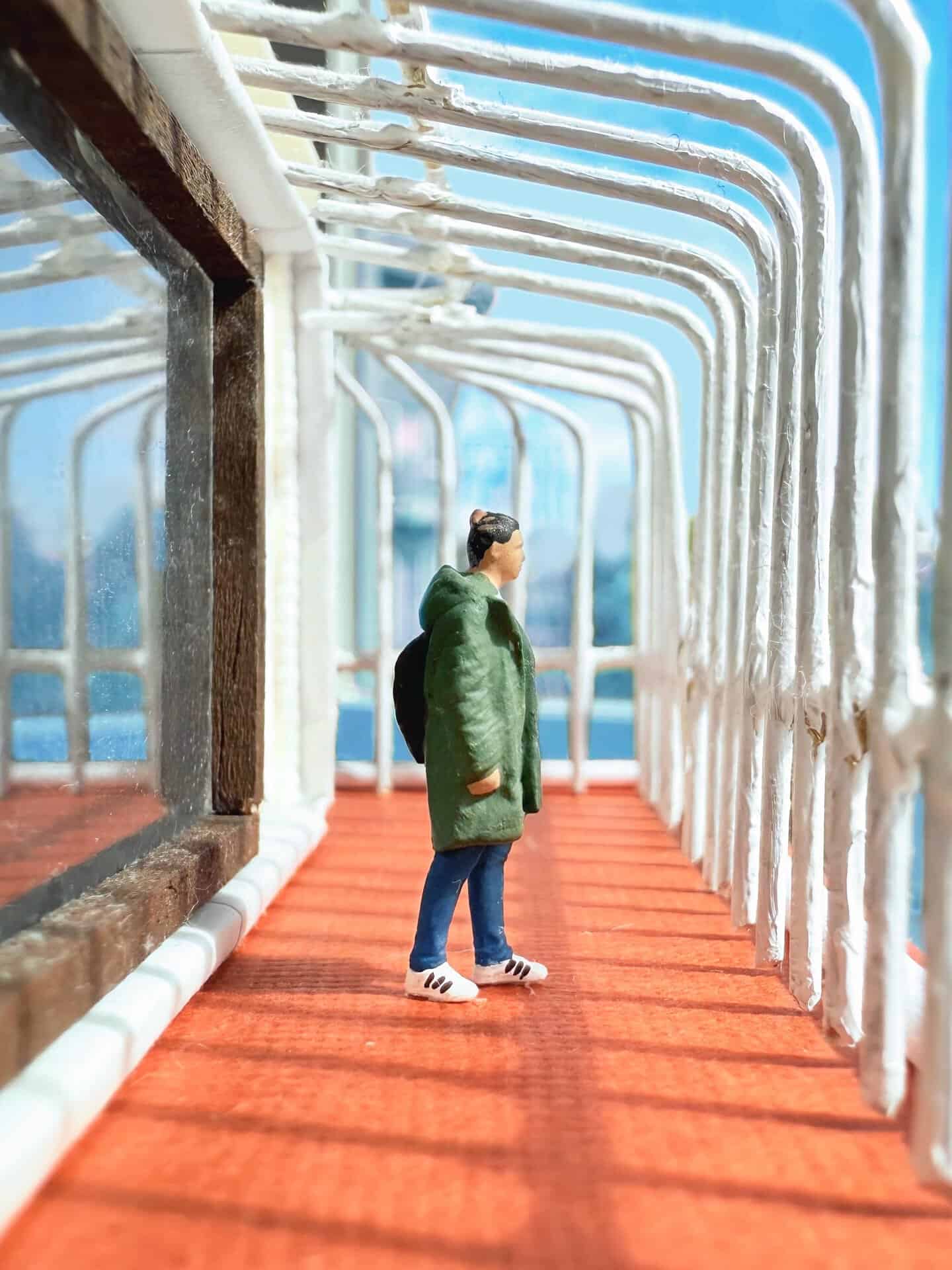 Miniature woman in green coat on orange walkway with white pipes overhead.