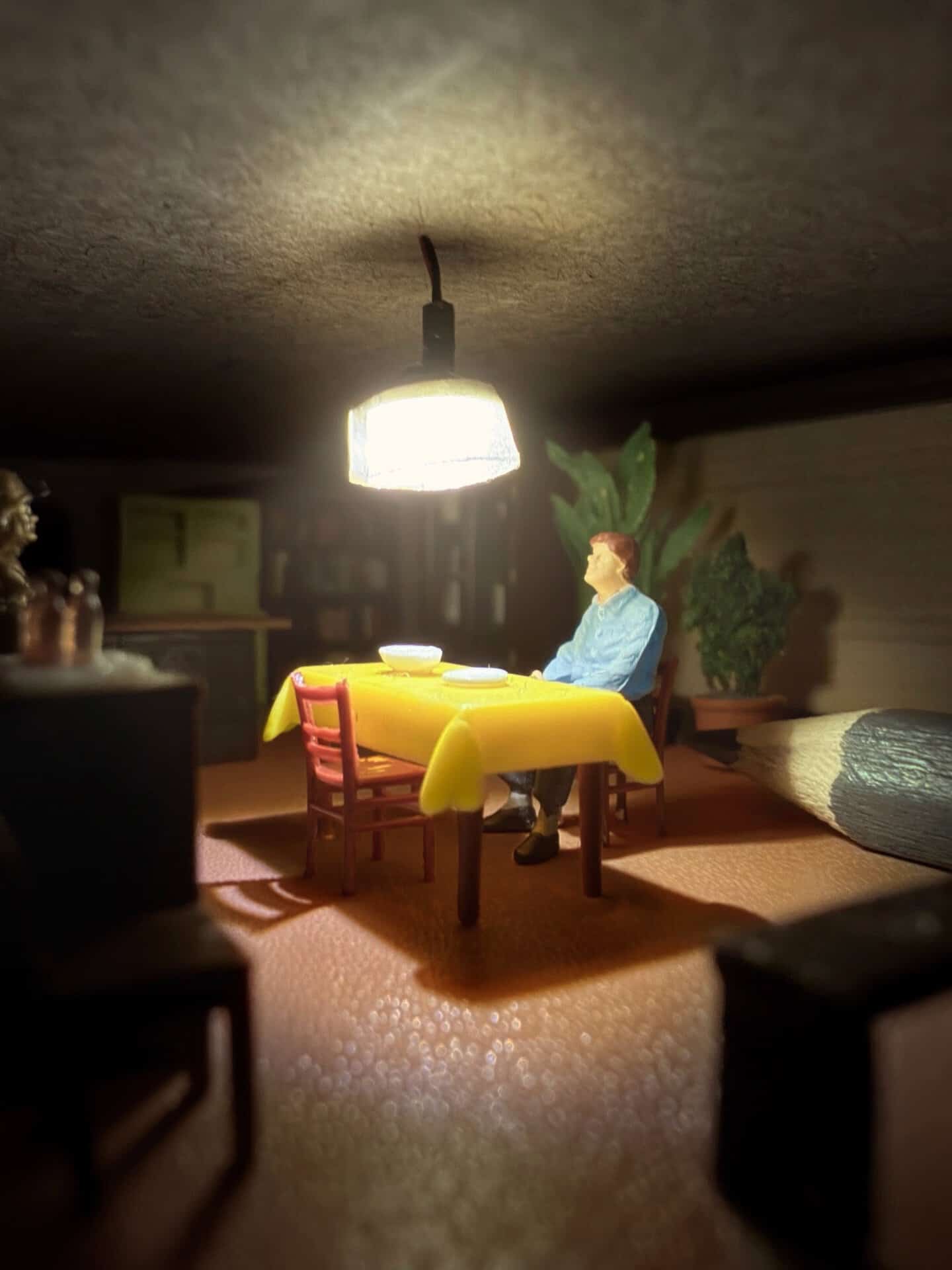 Miniature figure sitting at a table under a hanging light in a diorama scene.