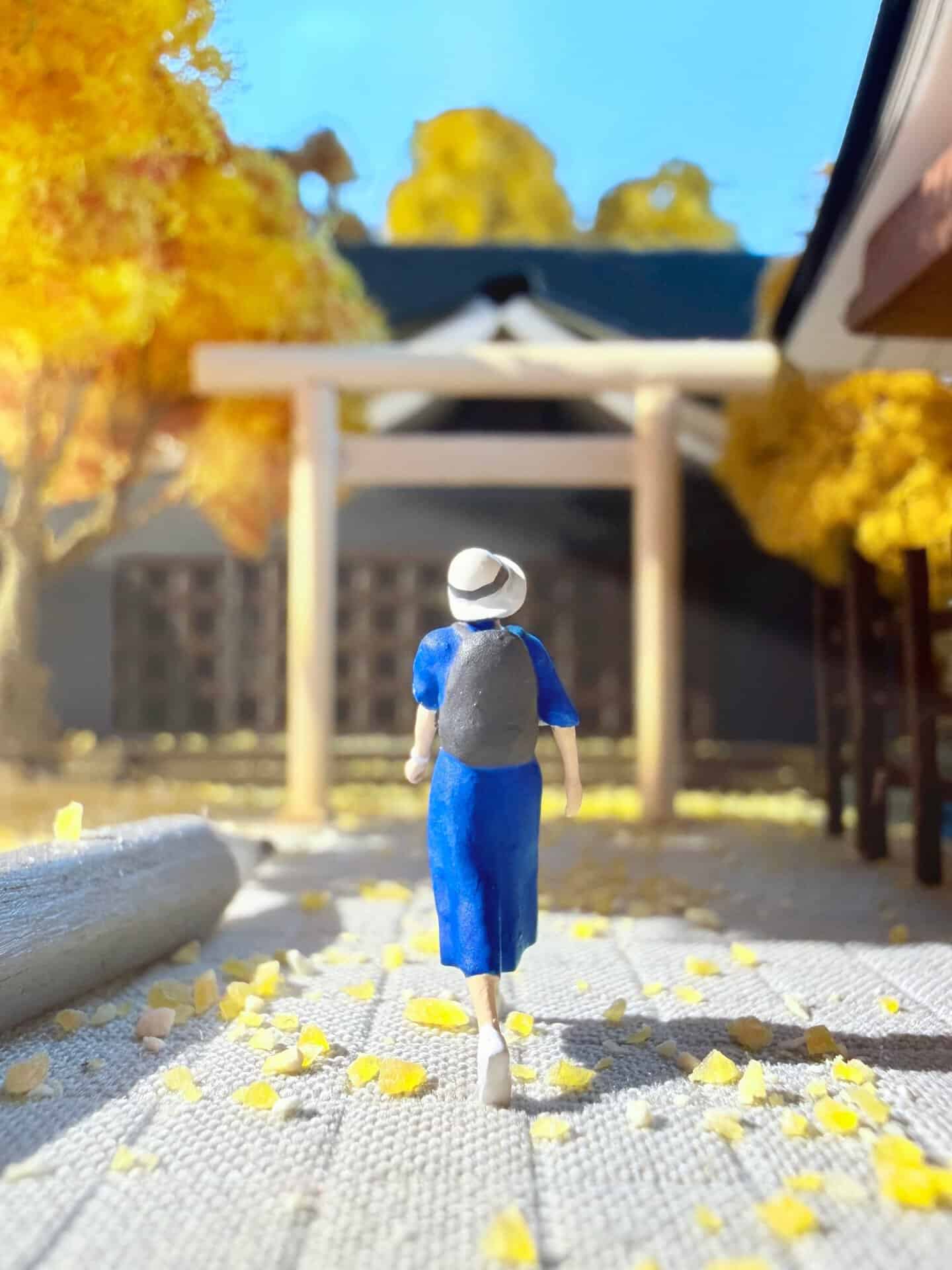 Miniature figure walking towards a traditional Japanese torii gate with autumn trees.