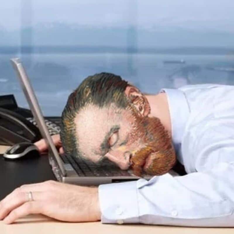 Man with painted face resting his head on a laptop at a desk.