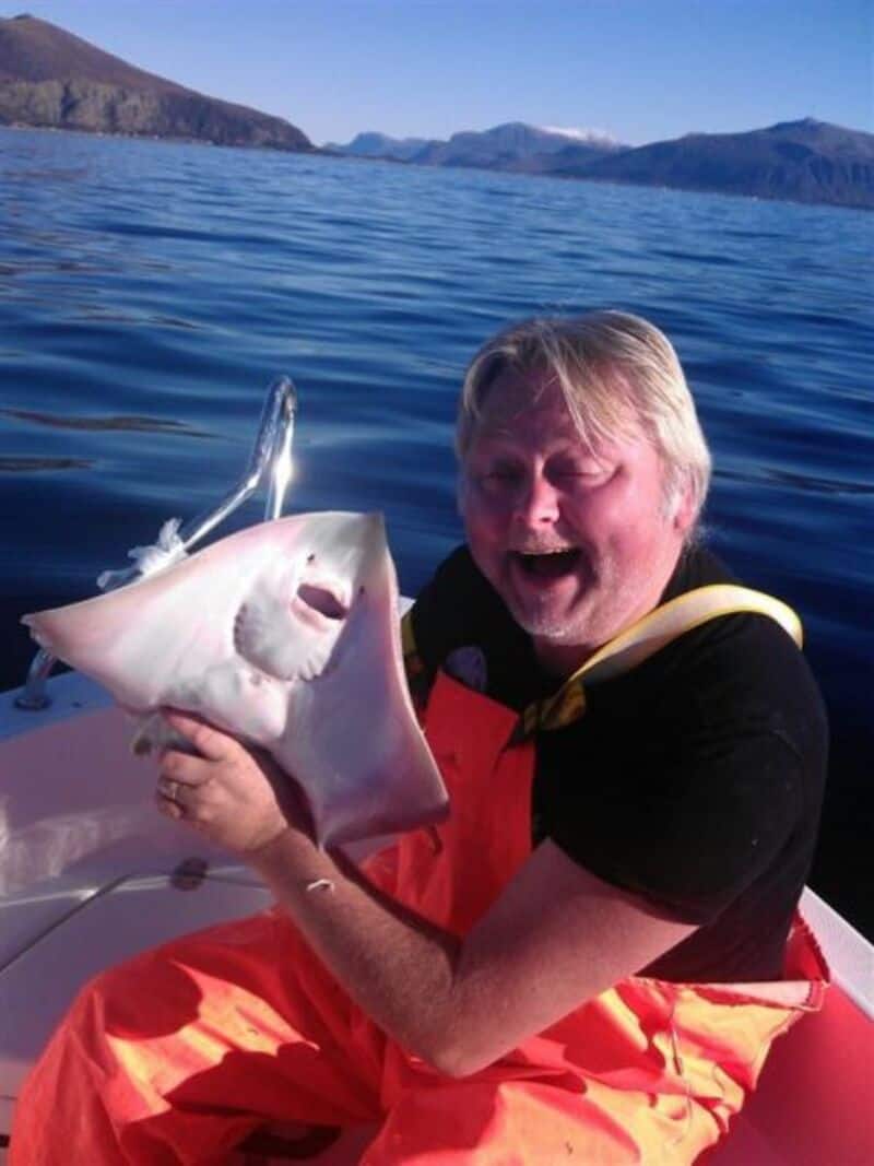 Man holding a large stingray on a boat with water and mountains in the background.