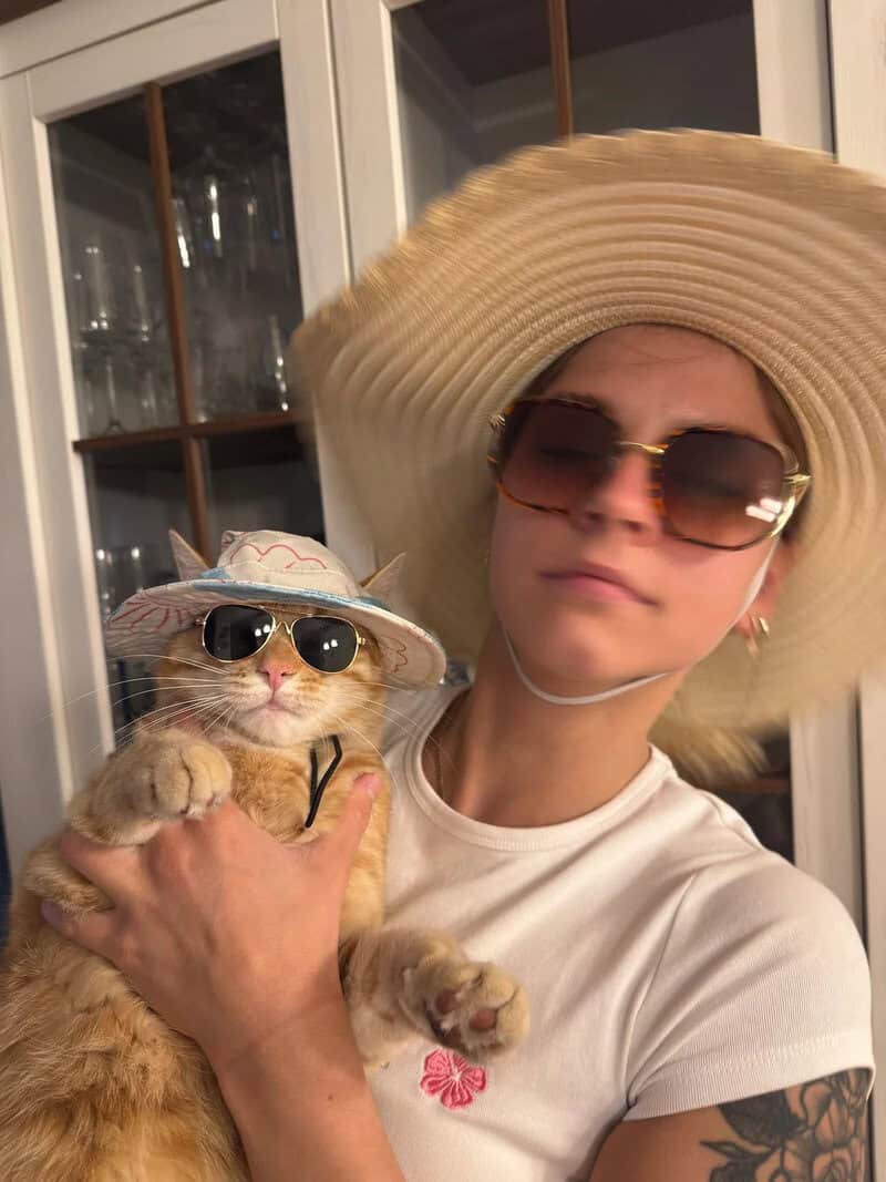 Person in large sunhat holding a cat with sunglasses and a hat.
