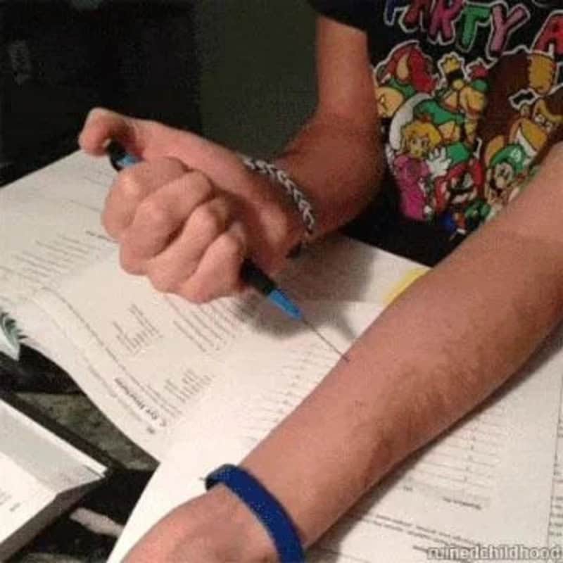 Person writing in a notebook with a blue pen, wearing a colorful cartoon T-shirt and a blue wristlet.