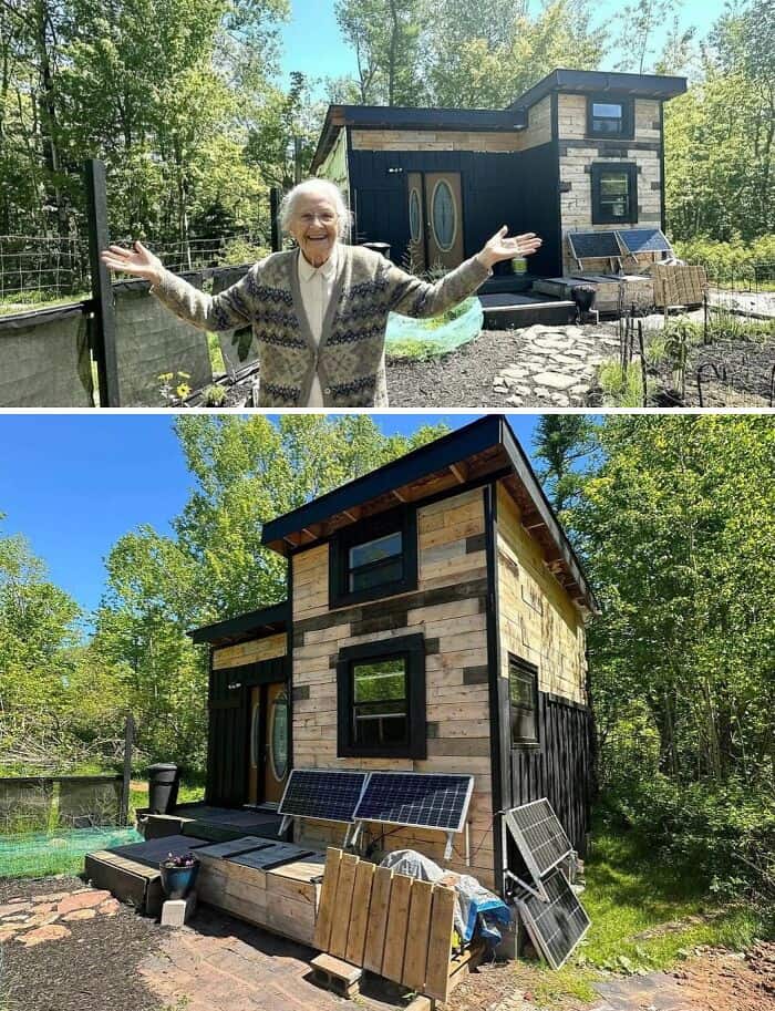 Woman smiling with arms open in front of a tiny modern house in a wooded area.