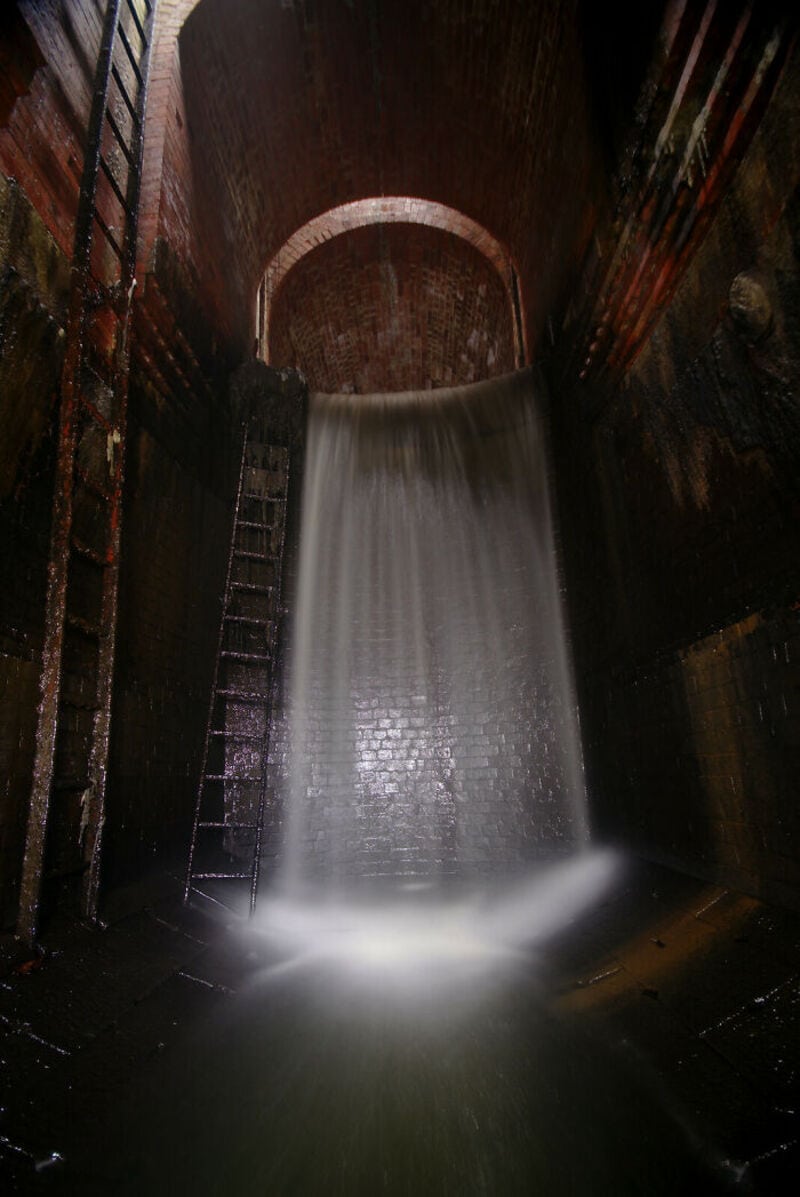 Water flows out of a large industrial pipe in a dark, brick-walled setting.