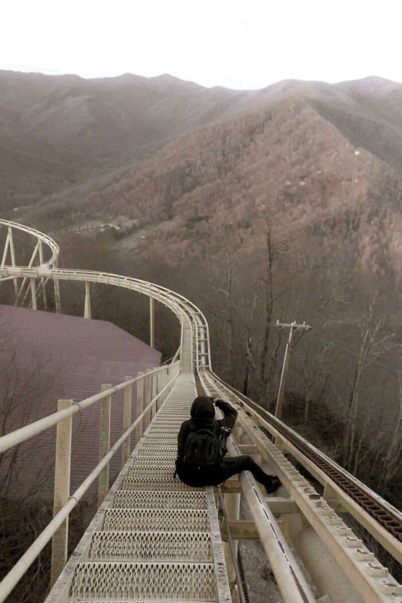 Person sitting on a roller coaster track overlooking mountains and trees.