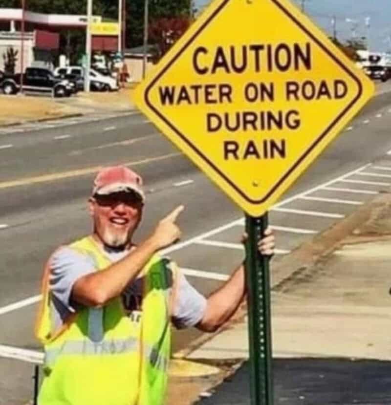 Man in yellow vest pointing at a caution sign about water on road during rain.