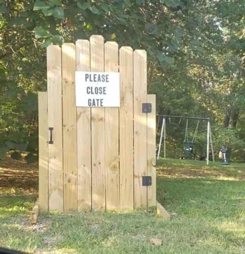 Please close gate sign on a wooden gate in a yard with trees and swings in the background.