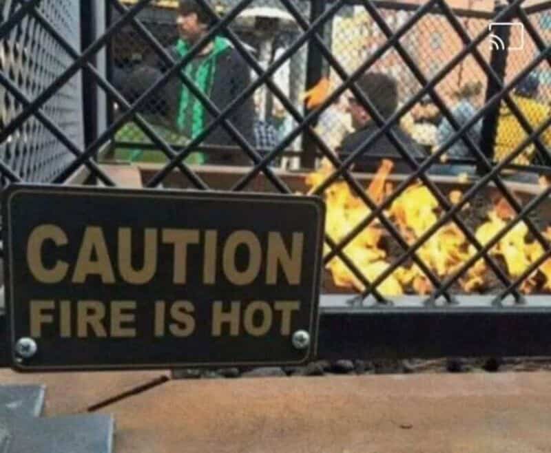 Caution fire is hot sign on a gate with people and yellow flowers behind.