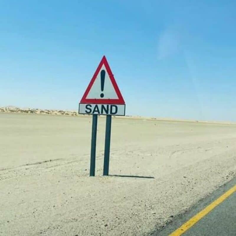Warning sign with exclamation mark and 'SAND' written below in a desert landscape.