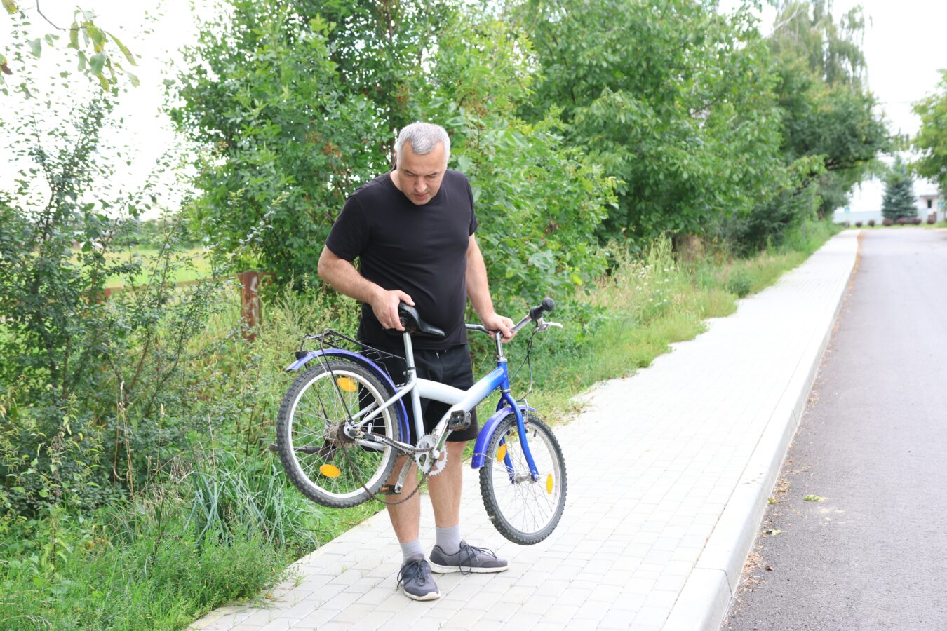 Older man fixing his bicycle on a sidewalk with greenery around.