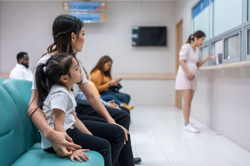 Children and adults waiting in a clinic, with a nurse at the window.