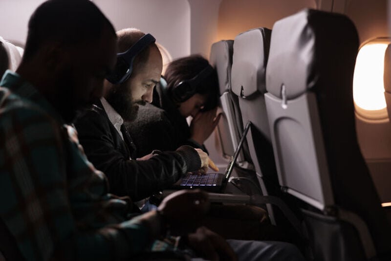Passengers on a plane wearing headphones, some using laptops or phones.