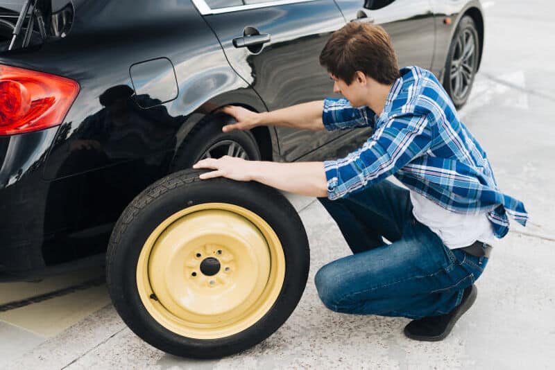Young man changing a flat tire on a black car in a parking lot.